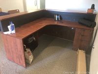 Front-left angle view of L-shaped office desk showing wood grain finish, built-in drawers, telephone, hand sanitizer, and open storage compartment under desk.