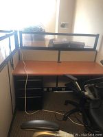 Cubicle desk with wood-tone surface, attached cubicle walls with black mesh and wood panel, and black three-drawer mobile pedestal under desk. Office chair partially visible.