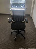 Black mesh office chair with armrests and caster wheels, positioned on a carpeted floor in front of a window.