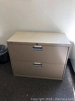 Beige 2-drawer filing cabinet with lock on top drawer, visible scratches and marks, placed against wall on carpeted floor.