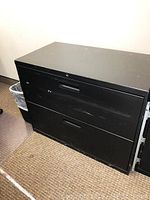 Black metal 2-drawer filing cabinet showing visible scratches and marks on surface. Positioned on carpeted floor with trash can beside.