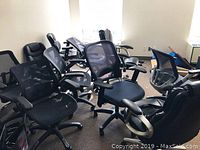 Ten black office desk chairs with mesh backs and fabric seats arranged in an office space near windows.