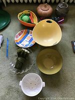 Top-down view of assorted serving bowls and decorative dishes on a carpeted surface including a basket bowl with faux vegetables, colorful patterned bowl, yellow bowls, white colander, and small lidded pot.