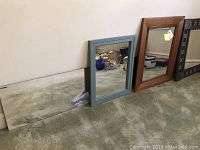 Four wall mirrors leaning against a wall on carpet; includes a large beveled mirror, a gray-blue framed mirror, a medium brown framed mirror, and a dark framed mirror with inlay
