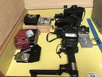 Photo showing assortment of digital and film cameras with boxes and cases on yellow table surface.