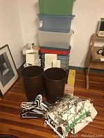 Various stacked plastic storage bins in green, blue, clear, and red with lids. Two large brown trash bins in front and multiple white and metal hangers scattered on the floor.