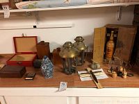 A collection of decorative pieces including ceramic ginger jar, two brass carriage lamp frames, multiple wooden boxes, a clothespin crucifix, and other miscellaneous items on a white shelf.