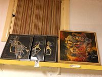Full view of four framed batik prints on yellow shelf showing three smaller traditional wayang prints and one larger colorful abstract print signed.