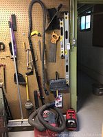 Photo of pegboard with assorted hand tools hanging including levels, hand saws, broom, plunger, crowbar, dustpan, ice scrapers, and wet/dry vacuum hose visible.