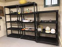 Three black plastic shelving units of different sizes against a basement wall with various items on shelves.