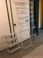 Photo showing a variety of white wire metal wall-mounted shelving units leaning against a door and wall. Shelves include angled wire shelves and basket-style wire shelves, tallest approximately 72 inches tall.