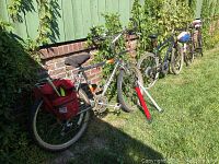 Three adult bicycles parked in grass next to a wall, featuring the Raleigh with a red pannier, Peugeot and Huffy mountain bike.