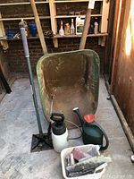 Large green metal wheelbarrow with hand tamper, sprayer, watering can, and white tub with concrete tools in front of a wooden shelf with garden chemicals.