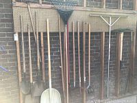Photo showing various garden rakes, shovels, and spades hung on pegboard against brick wall.
