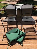 Two black metal mesh outdoor chairs shown from front, accompanied by two green seat cushions on the ground.