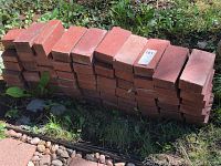 Stack of over 50 red standard size bricks outdoors on grass and stone.