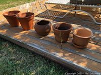 Six terra cotta plant pots arranged in a row on wooden deck, varying shapes and sizes.