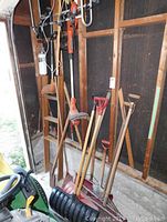 Two metal wall frames mounted vertically, holding two grass trimmers (one electric, one battery operated missing battery) and multiple wooden-handled shovels and rakes leaning against the wall.