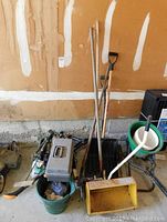 Photo showing various garden tools like long handled shovels and pruners, a green bucket with assorted tools, and a gray toolbox containing hardware.