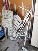 Photo showing white metal grocery cart, white resin lounge chair, dismantled metal crib parts, and a religious sign leaned against wall.