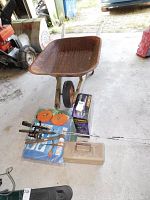 Wide view of rusty metal wheelbarrow, two fishing rods, boxed insect zapper, two tarps, small fishing tackle box on concrete floor indoors.