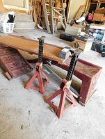 Two red Mastercraft 2 ton axle stands next to rusted metal car ramps and wooden creeper platform in garage.