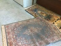 Two area rugs laid out on carpeted floor under sofa. Larger rug is blue with floral pattern showing wear and discoloration. Smaller rug behind has green and gold tones with fringed edges.