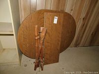 Oval dining table top, one rectangular leaf, and four detached turned legs tied together on a floor against wood-paneled wall.
