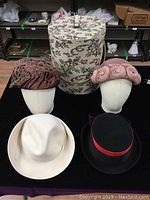 All four vintage hats positioned on mannequin heads with the floral patterned cylindrical hatbox behind them.