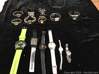 Photo showing 15 quartz watches arranged on a dark surface, featuring various styles and strap materials including metal, leather, silicone, and plastic. Bright green silicone band watch and digital calculator watch are visible.