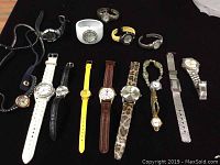 Full view of fifteen assorted quartz watches on black surface, various types and bands shown.