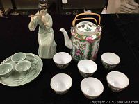 Photo of teapot with six matching cups, figurine, and tray with cups on black cloth surface
