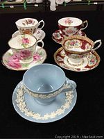 Front view of all five bone china tea cups and saucers arranged on black surface showing floral patterns and embossed relief on Wedgwood piece.