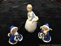 Three figurines on black fabric background: a Rex Valencia Spain 6-inch figurine of a girl with lamb and two Goebel West Germany angel figurines in blue robes holding lanterns.