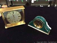 Two clocks on black cloth: Bulova quartz clock with rectangular case, gold-tone metal frame, and Roman numerals; Howard Miller mantel clock with green marble-like finish and gold trim, round dial, detached back