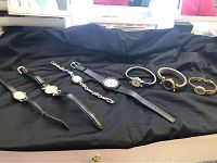 Photo showing seven costume wristwatches arranged in a row on black fabric, displaying various styles and band types.