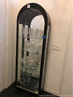 Full view of Philip Reinish curio cabinet showing arched top, black frame with gold trim, illuminated interior with glass shelves filled with crystal glassware.