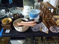 Wide view of kitchenware lot including coffee grinder, teapot, glassware, casseroles, wine rack, napkin holder, and wooden utensils arranged on a table.