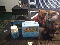 Snoopy and Woodstock musical piano in original box, Giorgio teddy bear, Smurfs lunchbox and thermos, and Caddyshack gopher figurine shown on table.