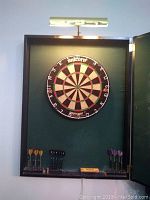 Dartboard and brass lamp mounted inside a custom green interior wooden case with darts stored below.