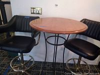 Photo showing round wood top pub table with metal pedestal base and two black upholstered barstools with metal bases.