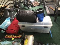Camping gear arranged on green carpet including black duffel bag, blue and black carry bag, folded tent and red bag, campfire grill box, and large plastic container.