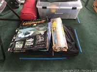 Photo showing camping equipment including a red carry bag, clear plastic storage bin, The Perfect Campfire Grill box, and other wrapped items arranged neatly on the floor.