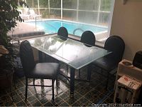 Glass dining table with four black upholstered chairs arranged around it beside a large window with pool view.