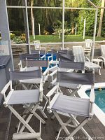 Eight white wooden folding directors chairs with grey canvas seats and backs arranged outdoors by poolside