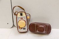 Front view of vintage Alfa camera with cream and red body next to its brown leather case.