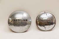 Two vintage metal hubcaps side by side: larger Studebaker hubcap on left, smaller Hupmobile hubcap on right, showing overall condition