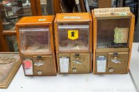 Front view of three vintage wood gumball vending machines side by side on a table, showing wooden cases, metal coin mechanisms, glass fronts, and pricing labels on two machines.