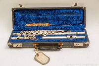 Open black hard case showing blue velvet interior with vintage Ambassador metal flute disassembled into three sections and cloth cleaning rod inside.