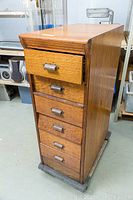 Six-drawer tall oak filing cabinet with visible wood grain and patina on metal pulls, standing on a dark base in a storage setting.